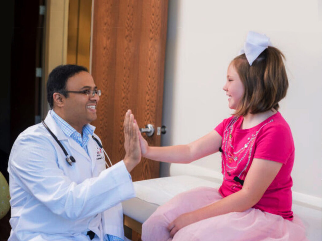 male physician smiling and giving high-five to child patient in pink shirt with white bow in hair, smiling