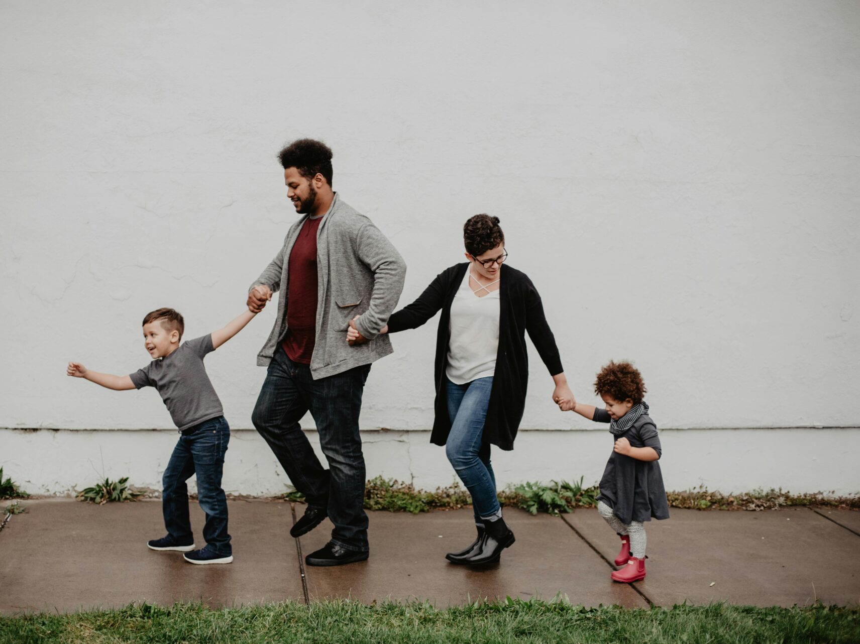 family with 2 kids walking on sidewalk in front of white wall, holding hands