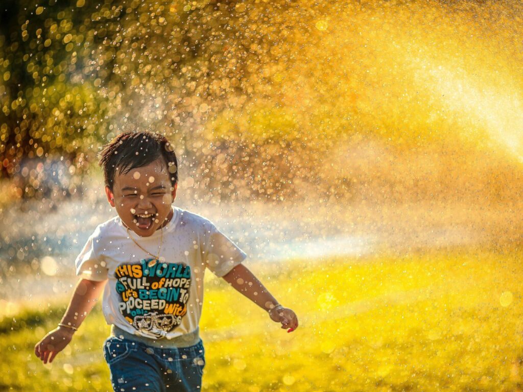 young boy running through water sprinkler, smiling with sun behind him