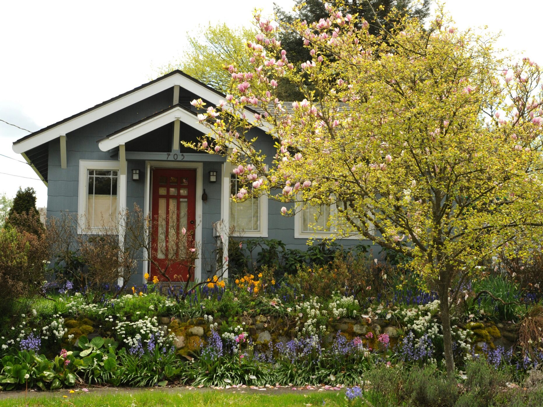 Exterior of residential home, painted gray with white trim with flowers and tree in foreground