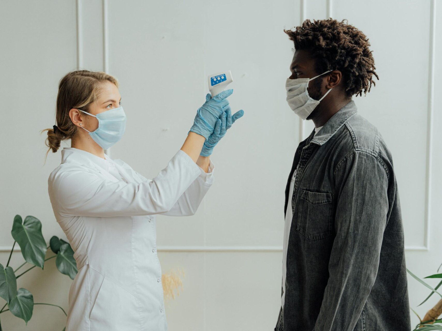 female doctor in white lab coat and blue gloves taking the temperature of a black male with mask