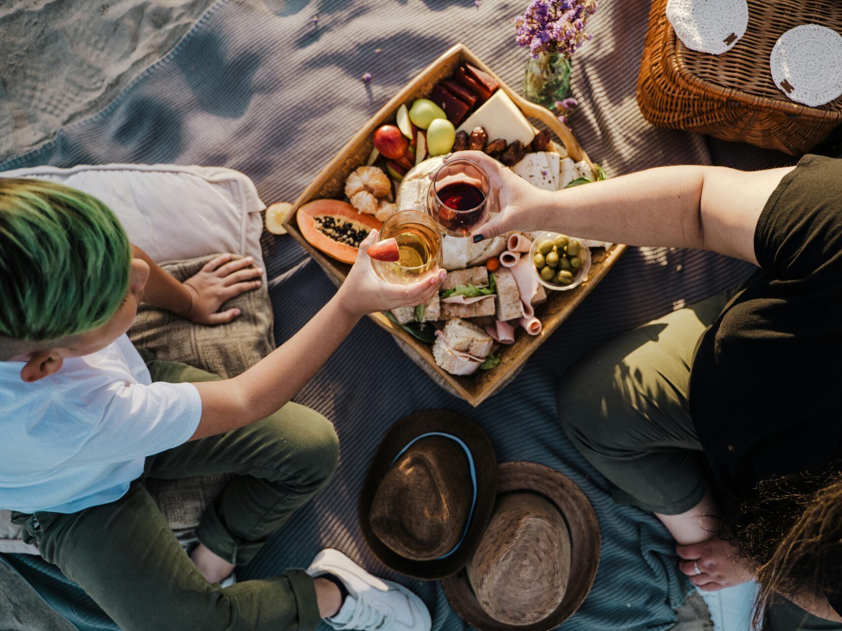 two people toasting glasses over charcuterie board on blanket during picnic