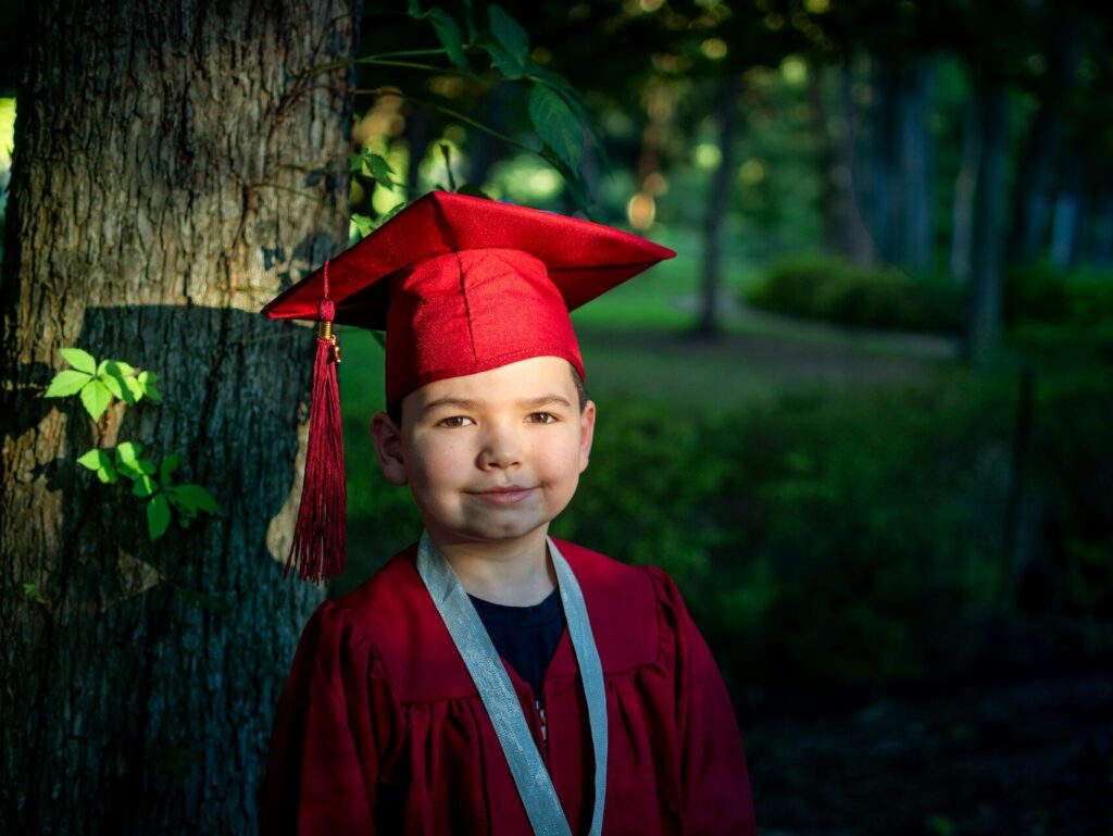 a male elementary student posing in front a free in cap and gown on graduation day