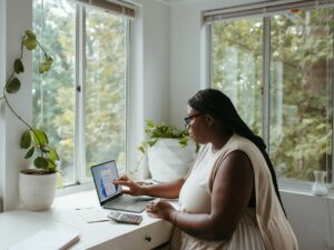woman sitting behind a white desk facing a window and working at her laptop