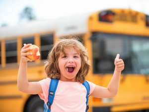 Young boy with backpack on holding an apple in one hand giving a thumbs up with the other and smiling in front of a school bus.