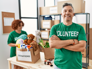 Man standing with arms crossed and green shirt that says volunteer standing in front of table with donated goods and woman sorting in background.