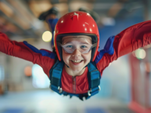 A kid with wind suit, harness, goggles, helmet who is facing camera with a huge smile on his face floating with arms out wide as he is indoor skydiving.