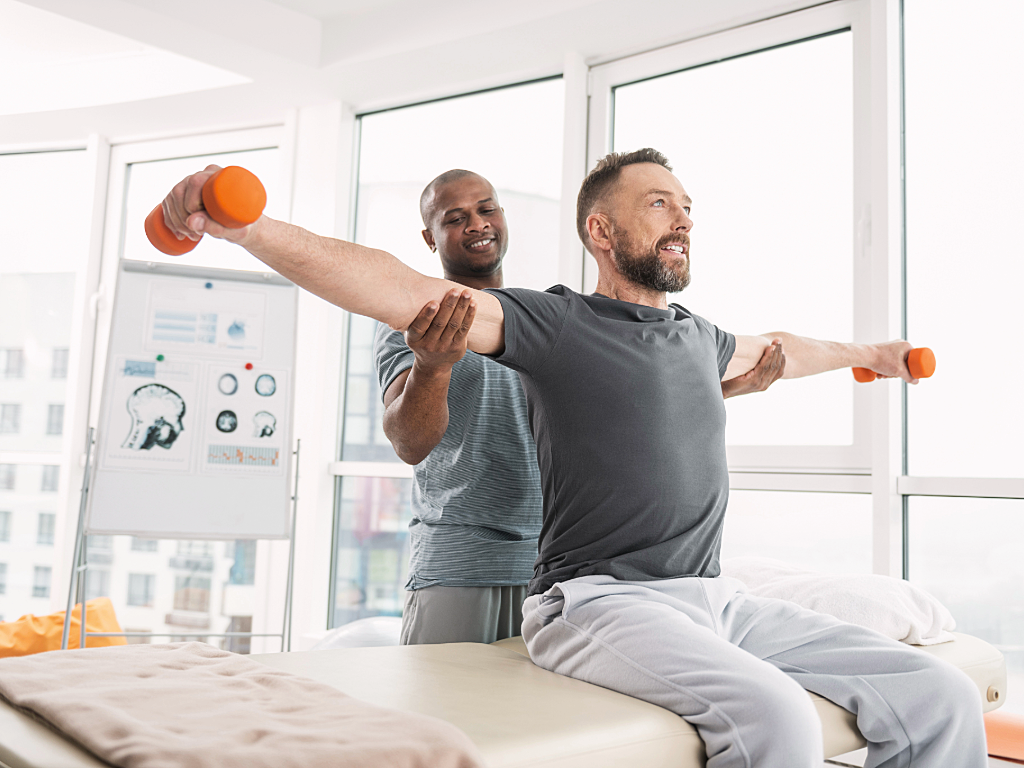 Physical therapist standing behind client helping to guide him as he raises arms holding small weights straight out to sides while sitting on table.