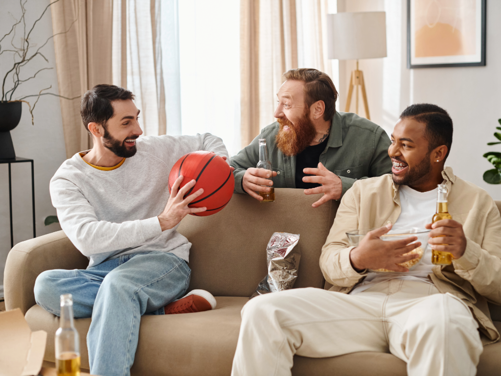 3 males - 2 sitting on couch, 1 standing behind the couch, drinking beer and watching a game together.