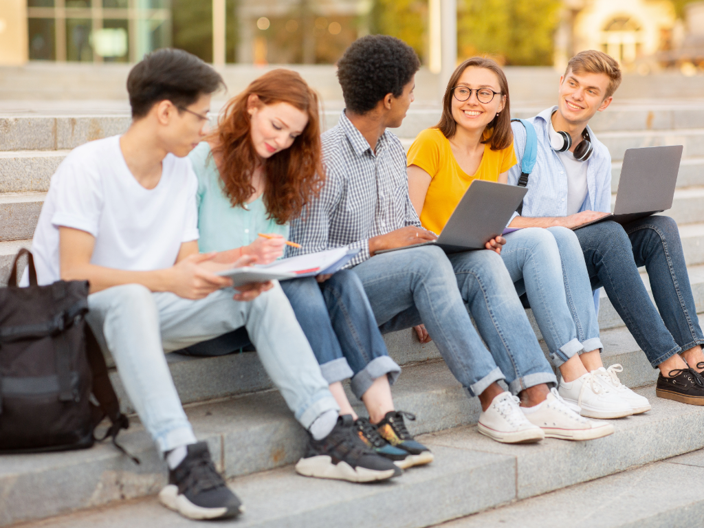 5 high school kids sitting on curb outside high school with books and laptops open.