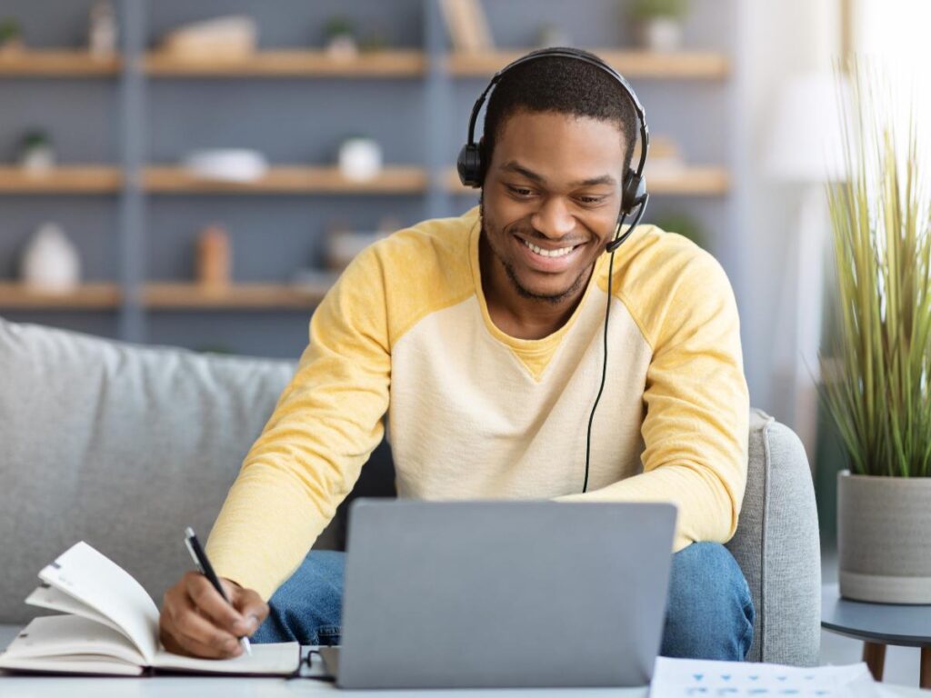 Black man sitting on couch smiling with headset on taking on online class on laptop and taking notes for article 7 Top Trade and Technical Schools in DFW for newcomers moving to DFW.