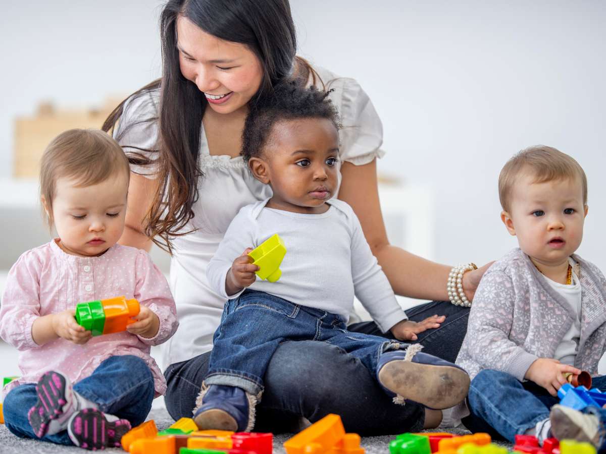 3 very young children with a woman smiling taking care of them as they play with blocks for article 2025 Ultimate List of Childcare Resources in DFW for newcomers moving to DFW.