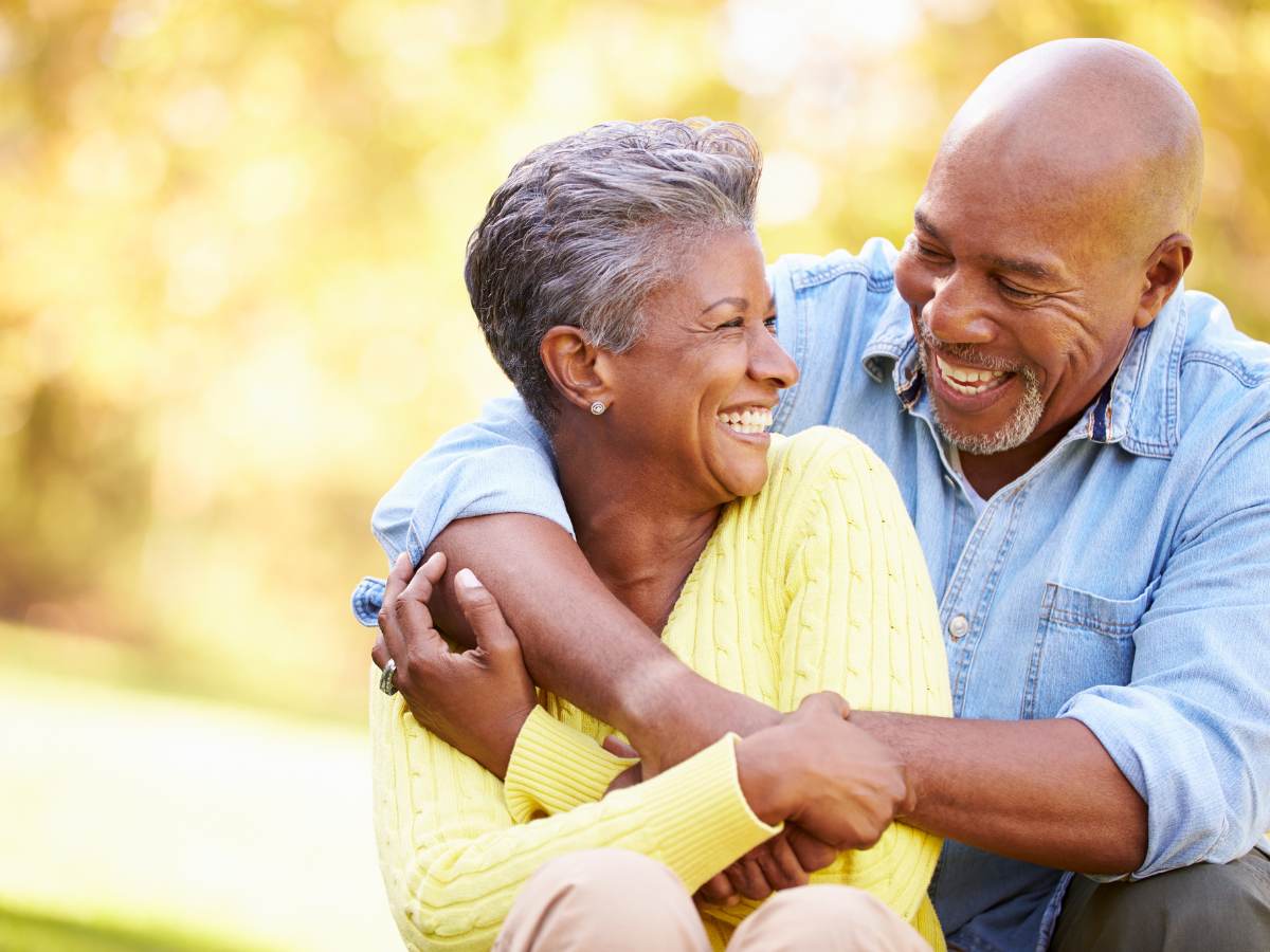 Senior adult Male with arms wrapped around senior adult female while sitting beside her. They are looking at each other and smiling. Bright yellow and green outdoor blurred background for article Senior Living in DFW for newcomers moving to DFW.