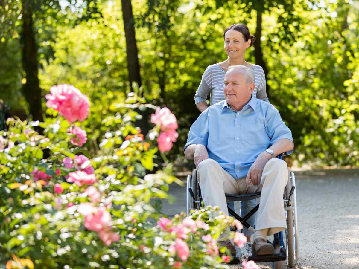 Senior woman pushing senior man in a wheelchair on a path through a nature area with trees in background and large pink flowers in foreground for article Retirement and Assisted Living in DFW for newcomers retiring to DFW.