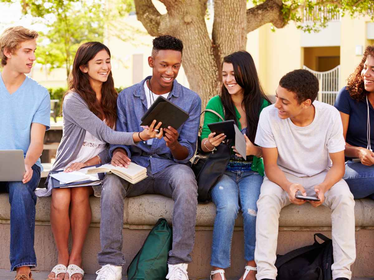 Group of 6 students sitting on concrete ledge holding tablets and with notebooks on their laps outside of school for article 20 Best Public High Schools in DFW for newcomers moving to DFW.