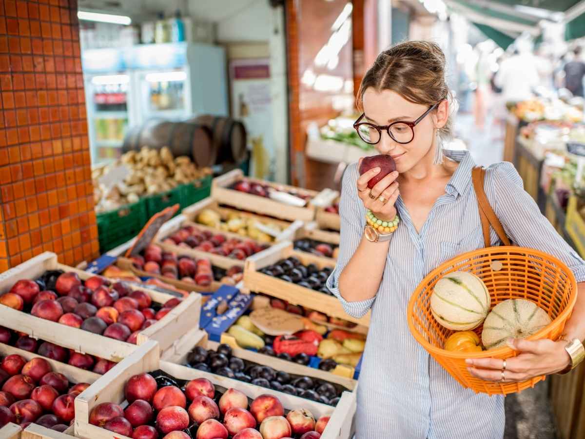 Woman walking through grocery aisle produce with basket of fruit and smelling an apple for article From Farm to Fridge: Where to Buy Organic Food in DFW for newcomers moving to DFW area.