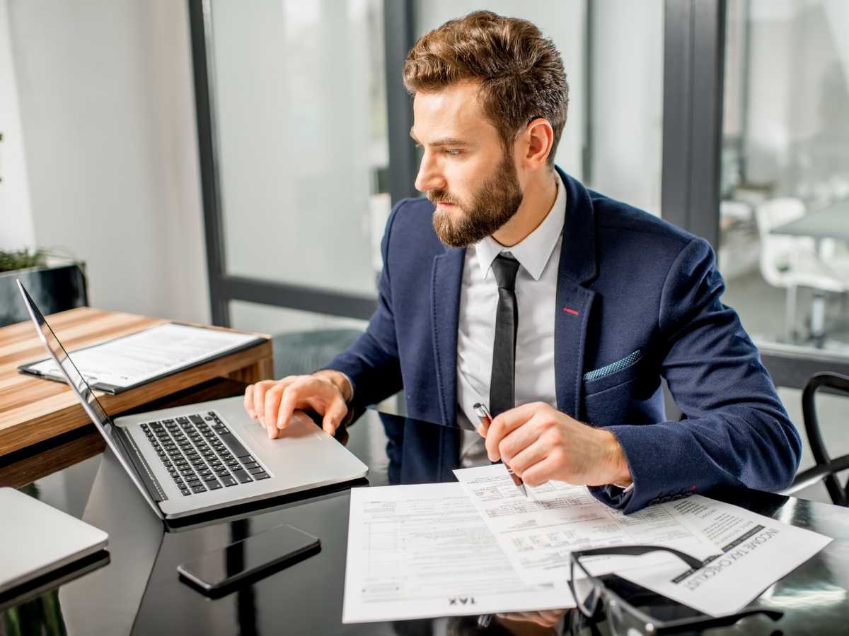Man in business suit sitting at desk in office working on laptop with tax forms around him for article No Income Tax! How That Shapes Your Budget in DFW