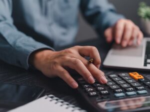 Close up of hand using a calculator on a desk with a laptop open for article Cost of Living in DFW vs Major U.S. Cities (2025 Comparison)